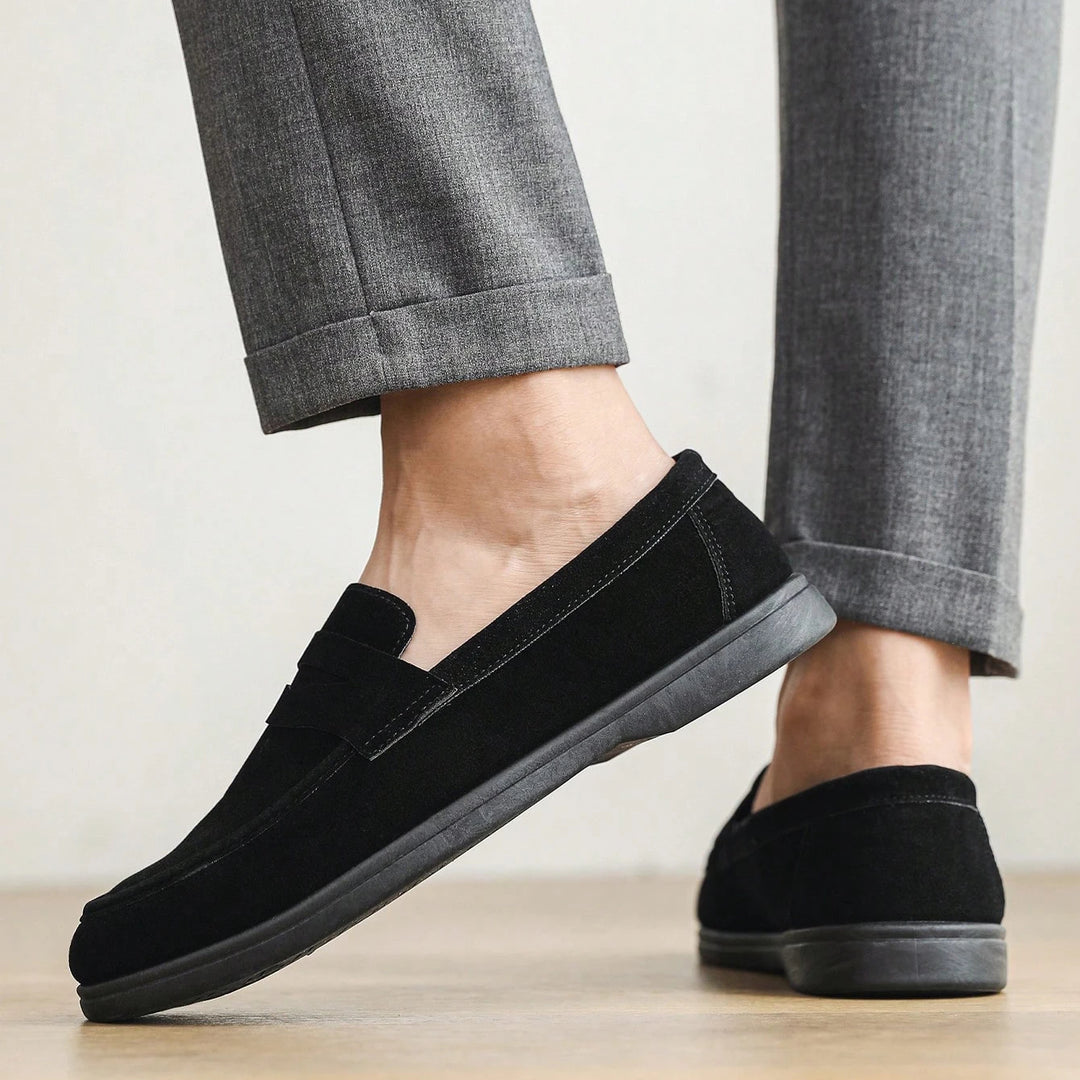 Black loafers worn with gray pants on a light wooden floor.