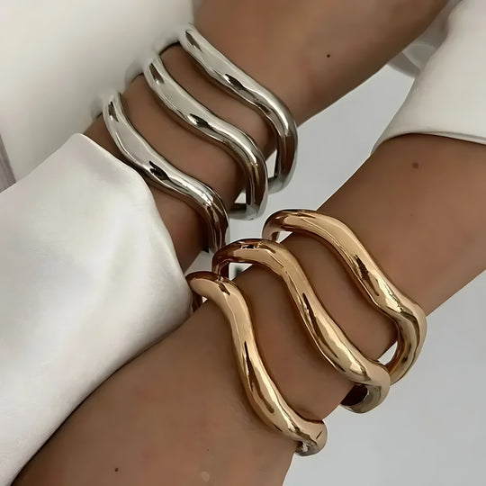 Two gold-plated metal bracelets with a bold wave design, displayed on wrists against a white background.
