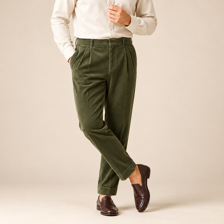 A person wearing olive green pleated trousers, a cream long-sleeve shirt, and dark brown leather loafers, standing against a plain beige studio background.