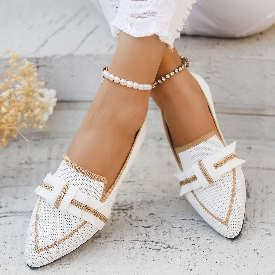 White pointed-toe shoes with gold accents worn by a person on a light wooden floor.