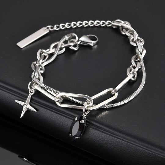 A silver chainlink charm bracelet with a polished snake and paperclip links, featuring a black gem and a cross charm, displayed on a black surface.