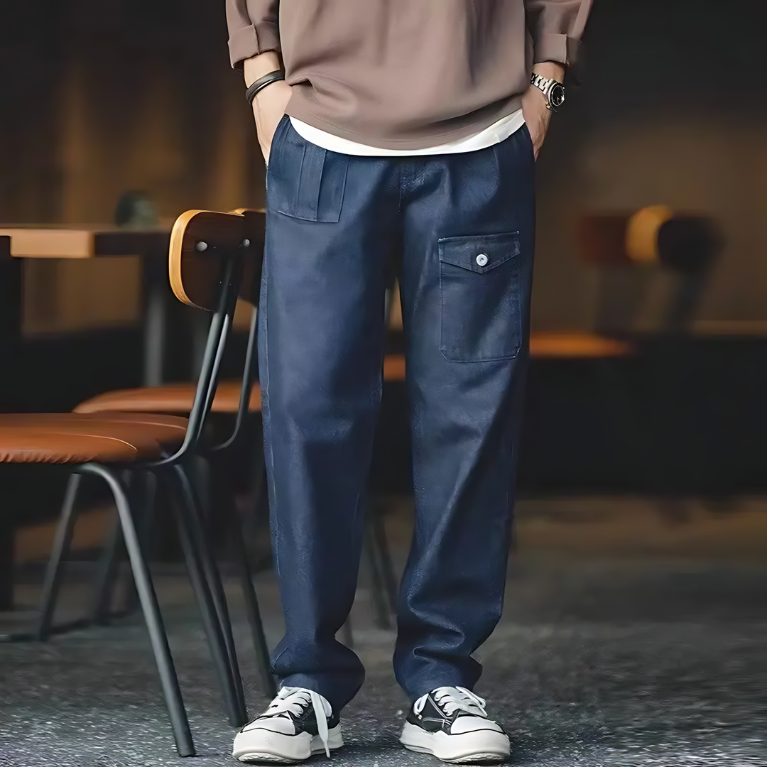 A person standing in a room with furniture, wearing loose-fitting blue denim jeans and a brown sweater, paired with sneakers.