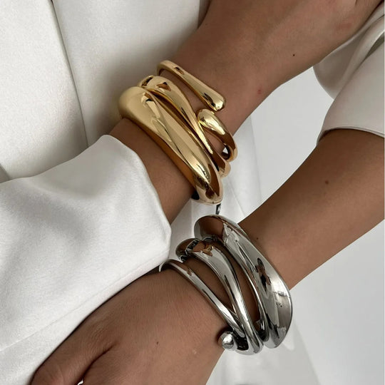 Two cuffs, one gold and one silver, with a fluid, wave-inspired design worn on a person's wrist.