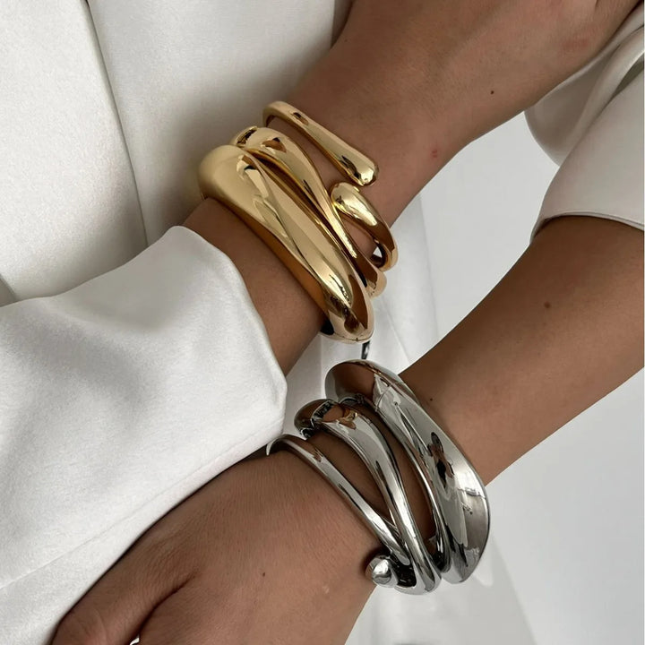 Two cuffs, one gold and one silver, with a fluid, wave-inspired design worn on a person's wrist.