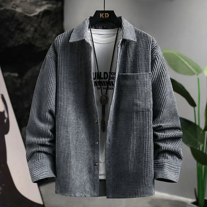 A beige corduroy shirt with a turn-down collar, long sleeves, and a single-breasted design. The shirt is buttoned at the top and displayed in a casual setting with plants and artwork in the background.