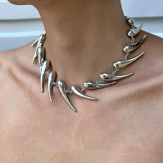 A close-up image of a stainless steel spike necklace worn around the neck, featuring a chain link design with multiple spikes protruding at different lengths.