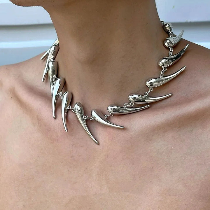 A close-up image of a stainless steel spike necklace worn around the neck, featuring a chain link design with multiple spikes protruding at different lengths.